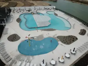 An aerial drone view of a large commercial gunite swimming pool featuring a zero-depth beach entry, a gated splash park with colorful water features, and an expansive concrete lounge deck.