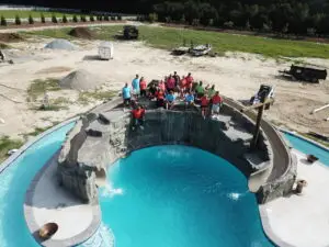 The Pools & Spas Unlimited (PSU) team posing for a group photo behind a massive custom gunite swimming pool featuring a large artificial rock grotto and a built-in water slide.