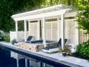 A luxury custom fiberglass spa with a raised stone veneer exterior, integrated into a pool deck under a white pergola with modern patio furniture.