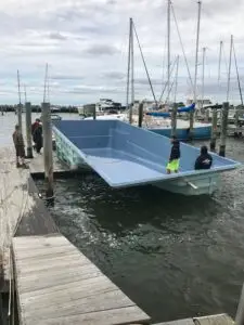 A light blue Latham fiberglass pool shell floating in the water and being towed by a ski boat down the Delaware Bay past a wooden dock and sailboats.