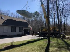 A large yellow crane lifting a white Latham fiberglass swimming pool shell over a residential house for installation in a backyard.