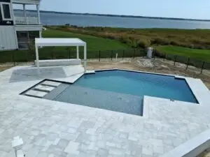 A rectangular geometric swimming pool with a light blue interior, featuring a tanning ledge and stepping stones, surrounded by a light grey stone paver patio overlooking a bay.
