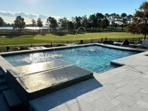 A rectangular gunite swimming pool featuring deck-mounted laminar jets arcing into the water, an integrated spa with a cover, and a light-grey stone paver patio overlooking a golf course.