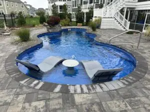 A freeform Latham fiberglass swimming pool with a vibrant blue finish, featuring an integrated tanning ledge with grey lounge chairs and a matching table, surrounded by a grey stone paver patio.