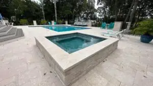 A custom square raised gunite spa with a light stone veneer, integrated into an expansive light-toned travertine tile patio next to a geometric swimming pool.