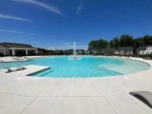 A large, circular commercial gunite swimming pool with a central tiered fountain, wide concrete deck, and residential community buildings in the background.
