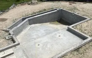 A residential gunite swimming pool under construction, showing the newly sprayed concrete shell with a deep end and integrated steps in a backyard setting.