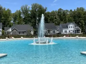 A large commercial gunite inground swimming pool featuring a decadent multi-tiered center fountain water feature, surrounded by a concrete patio and residential buildings.