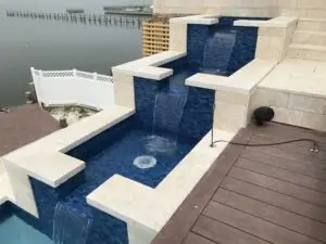 A luxury custom gunite spillover water feature fully tiled in blue glass mosaic, integrated into a multi-level stone patio with a dark wood deck.