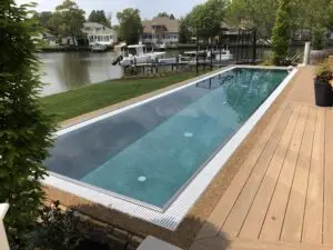 A luxury rectangular gunite lap pool with a vanishing edge overlooking a bay, featuring a light-colored wood deck and professional landscaping.