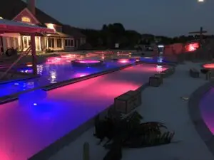 A nighttime view of a massive residential gunite pool with vibrant purple and pink LED lighting, featuring a 300-foot lazy river, an elevated spillover spa, and a custom rock waterfall.