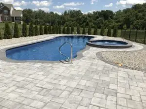 A luxury fiberglass inground swimming pool with a matching fiberglass spillover spa, surrounded by a large grey and tan stone paver patio and a black aluminum safety fence.