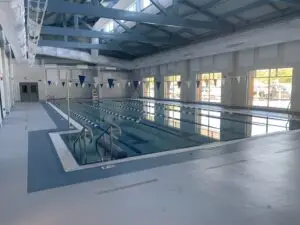 A large commercial gunite indoor swimming pool located inside a school facility, featuring multiple swim lanes, a built-in access ramp with railings, and large windows for natural light.