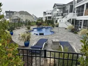 A luxury fiberglass inground swimming pool with a sun shelf and integrated steps, surrounded by a large grey stone paver patio and professional landscaping in a residential backyard.