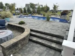 A luxury fiberglass inground swimming pool with a built-in spa, surrounded by a multi-level stone paver patio, masonry retaining walls, and professional landscaping.