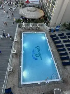 An overhead view of a commercial gunite swimming pool located on a bustling boardwalk, featuring a large "Sands" logo on the pool floor, surrounded by lounge chairs and a tiki bar area.