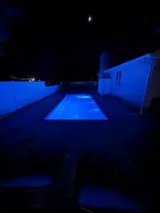 A rectangular fiberglass inground swimming pool at night, illuminated by vibrant blue Hayward 320 LED lights, situated next to a residential home with a white privacy fence.