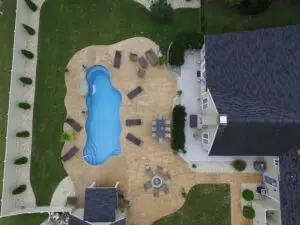 An aerial drone view of a custom Latham fiberglass inground swimming pool with a freeform design, featuring a tan stone paver patio and multiple outdoor seating areas.